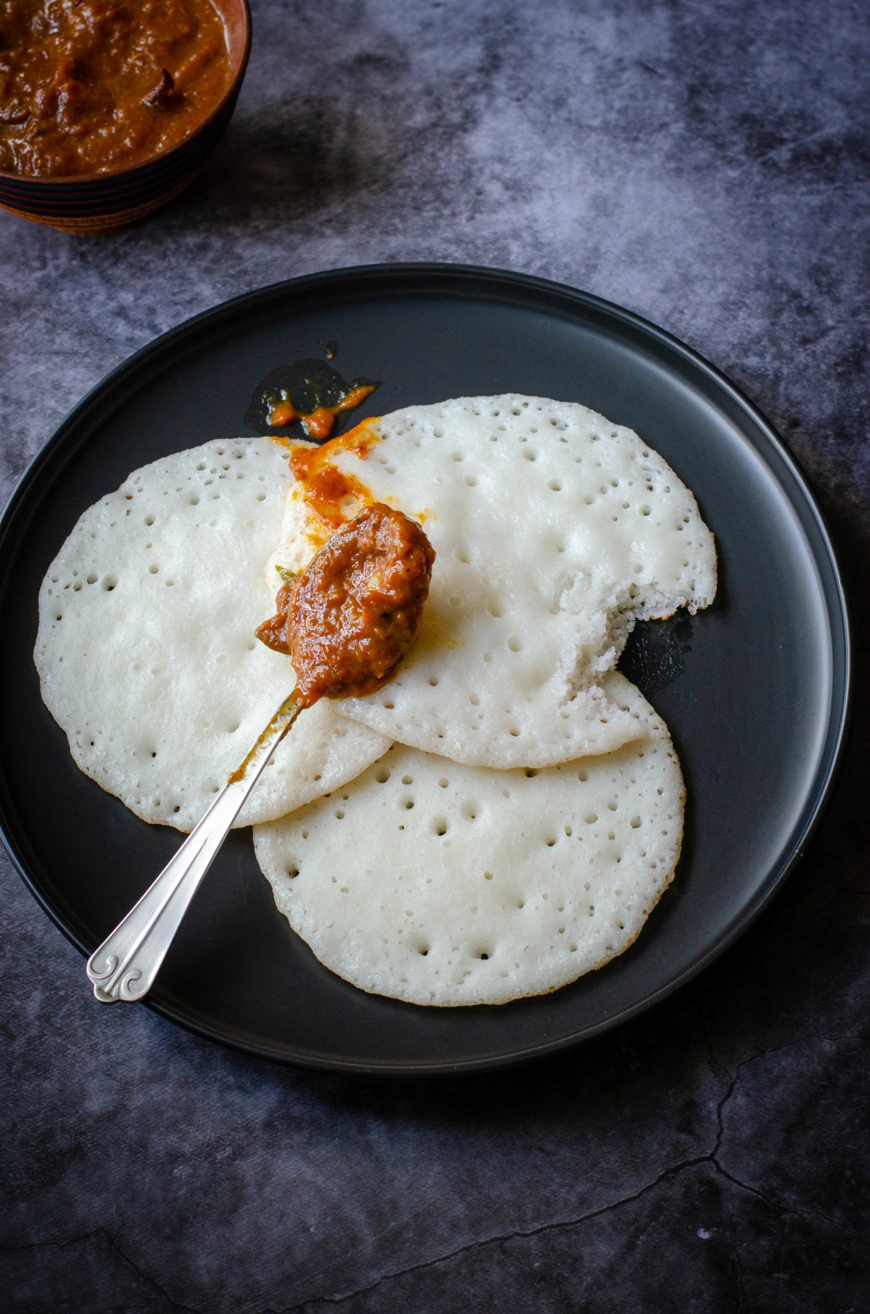 no ferment sponge dosa & tomato thokku served on a black plate set against a grey backdrop