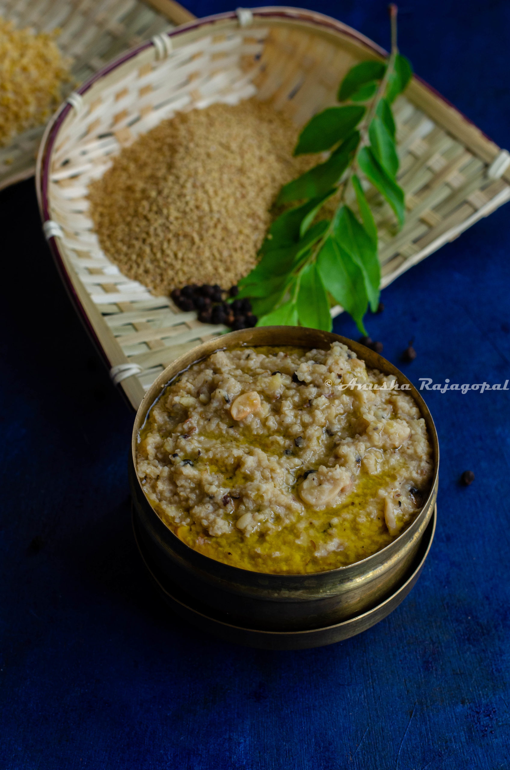 Thinai Ven Pongal served in a brass box with ghee on the top. Curry leaves and millet in a winnow as a backdrop.