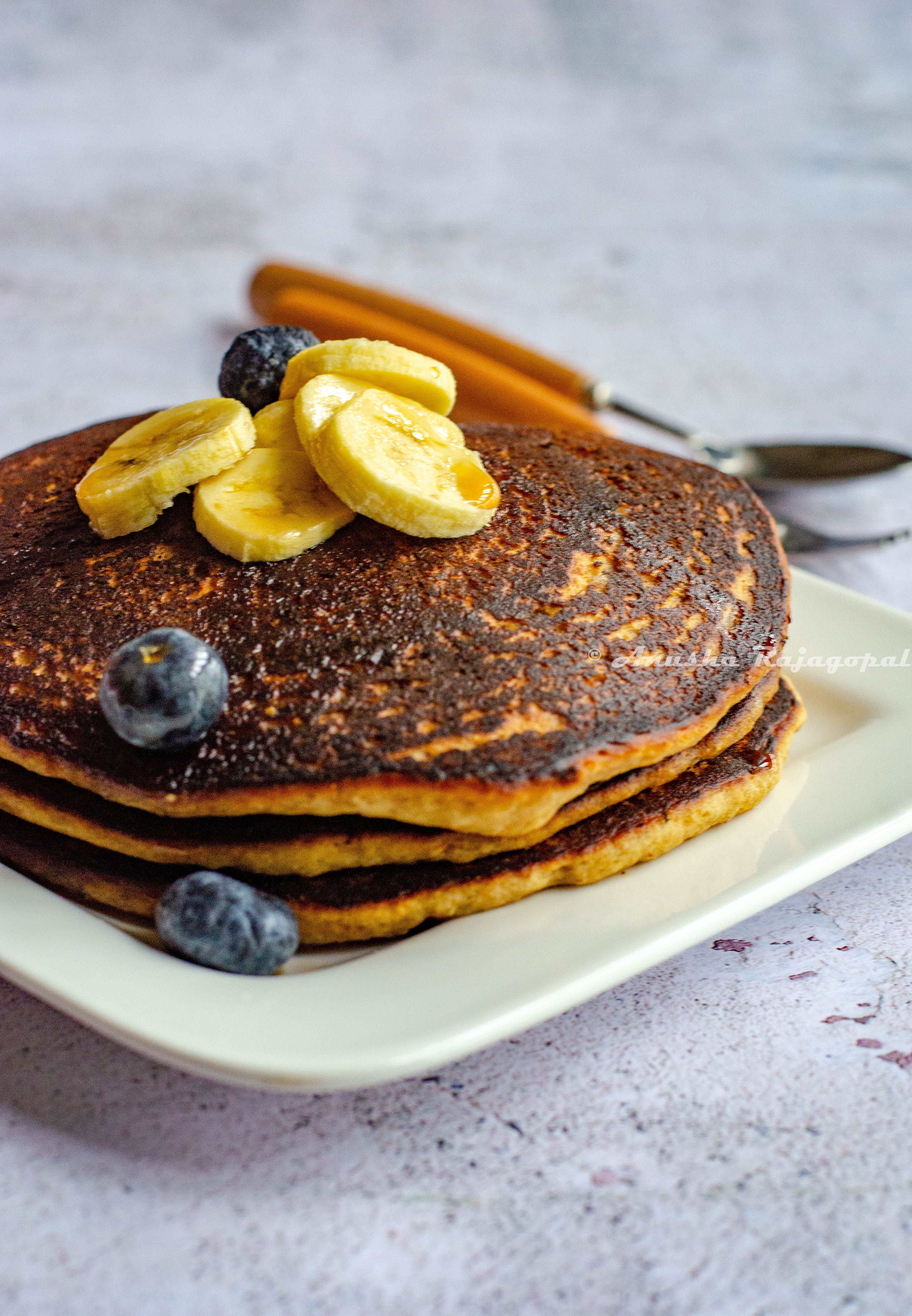 vegan banana oatmeal pancakes stacked and served on a white square plate. Banana and blueberries as toppings