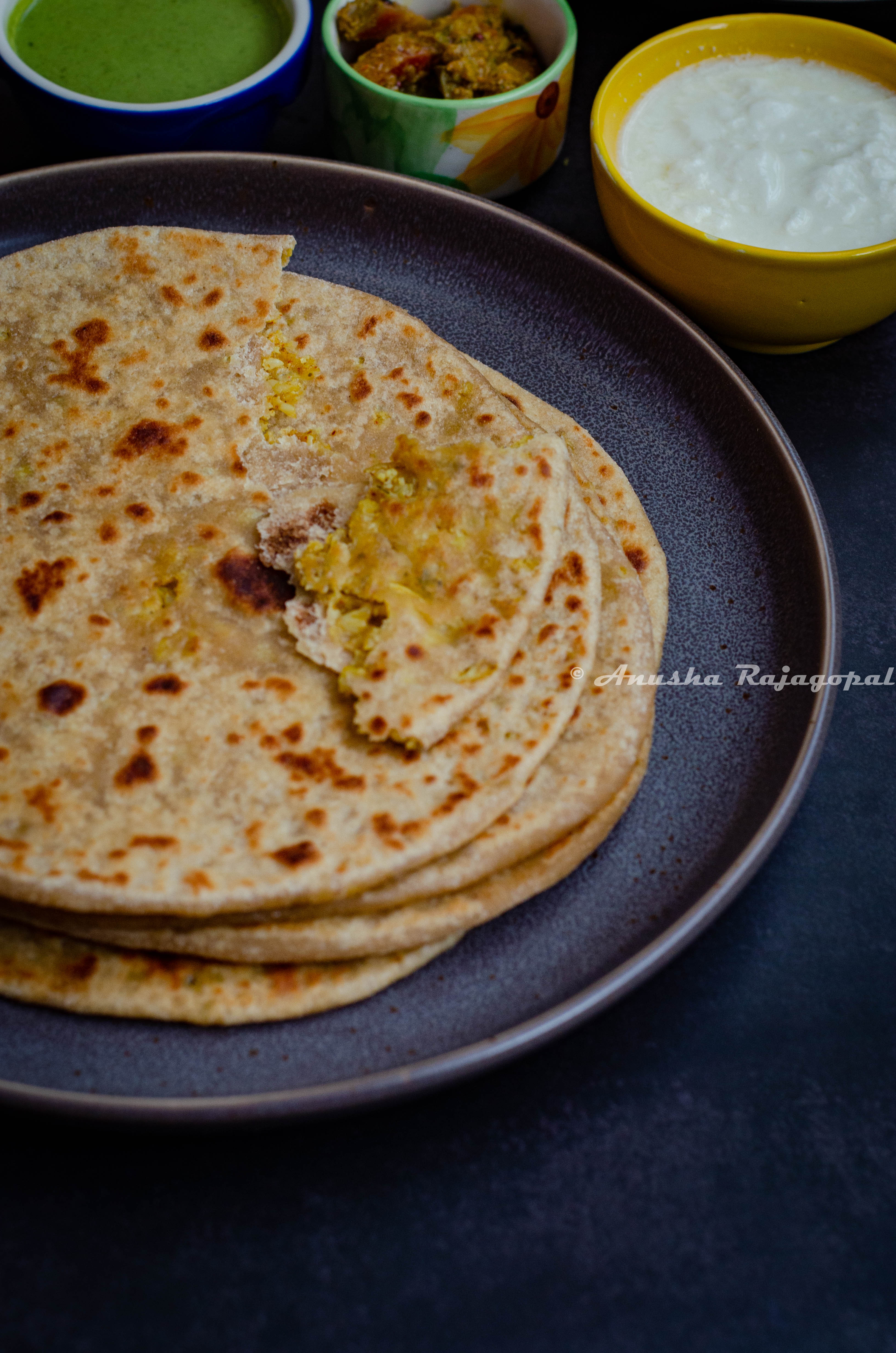 Gobi Paratha, Indian style cauliflower stuffed flatbreads stacked and served on a black plate. Yogurt and pickles at the back.